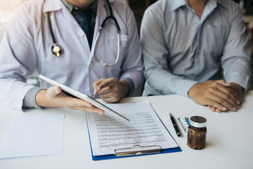 A doctor in a white coat showing a digital tablet to a patient at a desk with a medical clipboard and a pill bottle.
