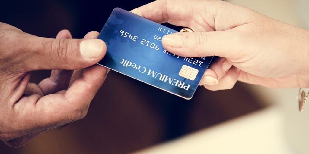 Close-up of a handoff where a person passes a blue prop credit card featuring an EMV chip and the text "Premium Credit" to a merchant.