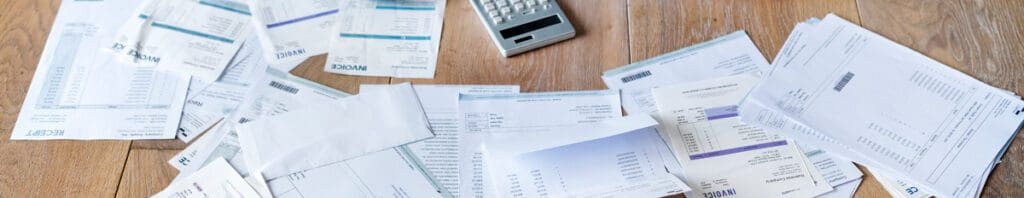 A cluttered mess of household bills, invoices, and papers spread across a wooden floor next to a silver calculator, symbolizing financial confusion and stress.